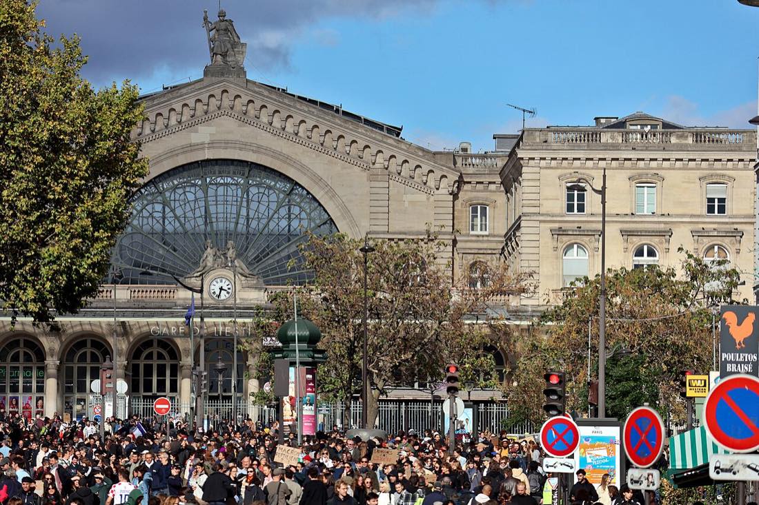 A large crowd of marchers in front of a Paris railway station with an arched glass facade.